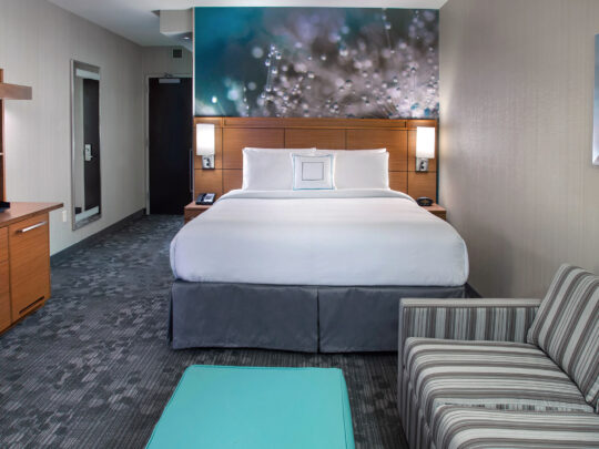 Um quarto de hotel no Courtyard South Philadelphia, no Navy Yard, com uma cama king-size branca e cinza, um sofá listrado, uma pequena mesa azul-petróleo e uma TV sobre uma cômoda.