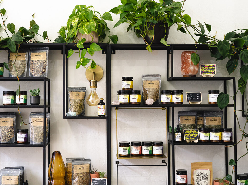 Teas, herbs, elixirs and powders are displayed on a black metal shelf against a white wall. Leafy green plants are on the top shelf.