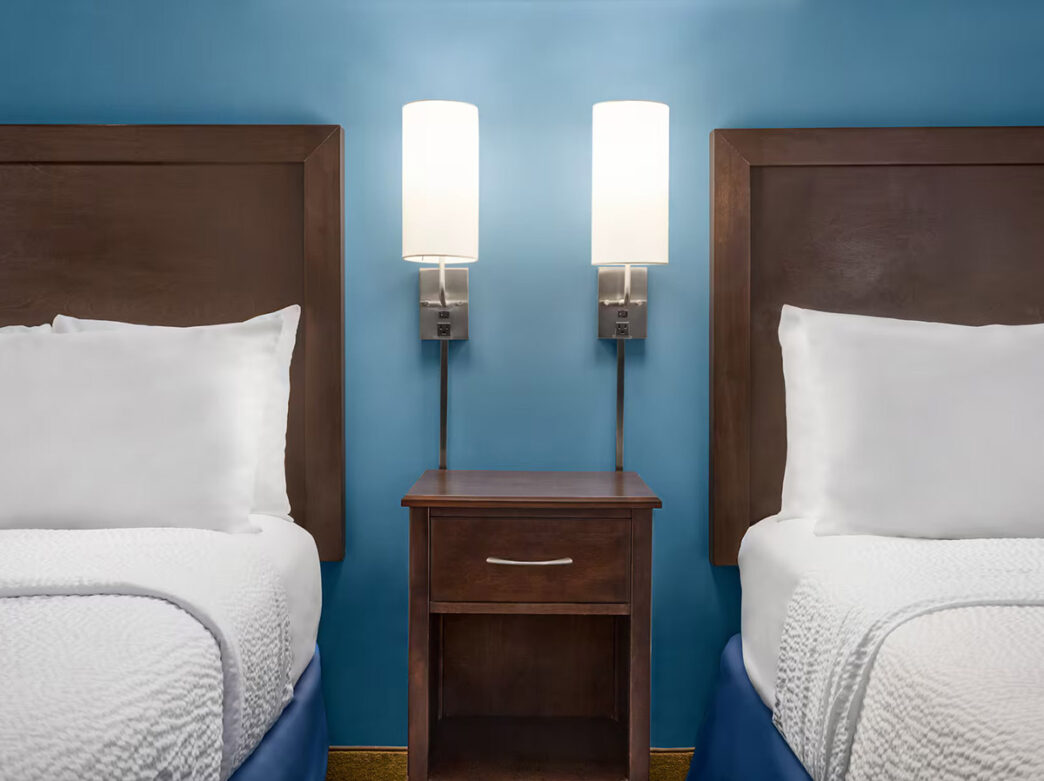 A hotel room inside the Days Inn by Wyndham Philadelphia Convention Center with two white beds, two white lights, a brown nightstand and light blue wall.