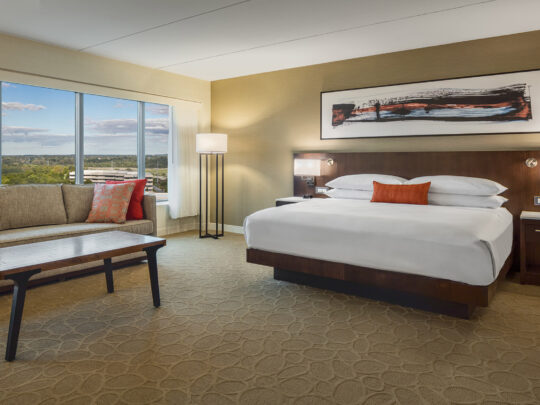 Moderna habitación de hotel con cama extragrande con sábanas blancas, zona de estar con sofá de felpa y mesa de centro, y ventanales con vistas panorámicas.