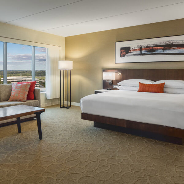 A modern hotel room featuring a king-size bed with crisp white linens, a seating area with a plush couch and coffee table, and floor-to-ceiling windows showcasing scenic views.