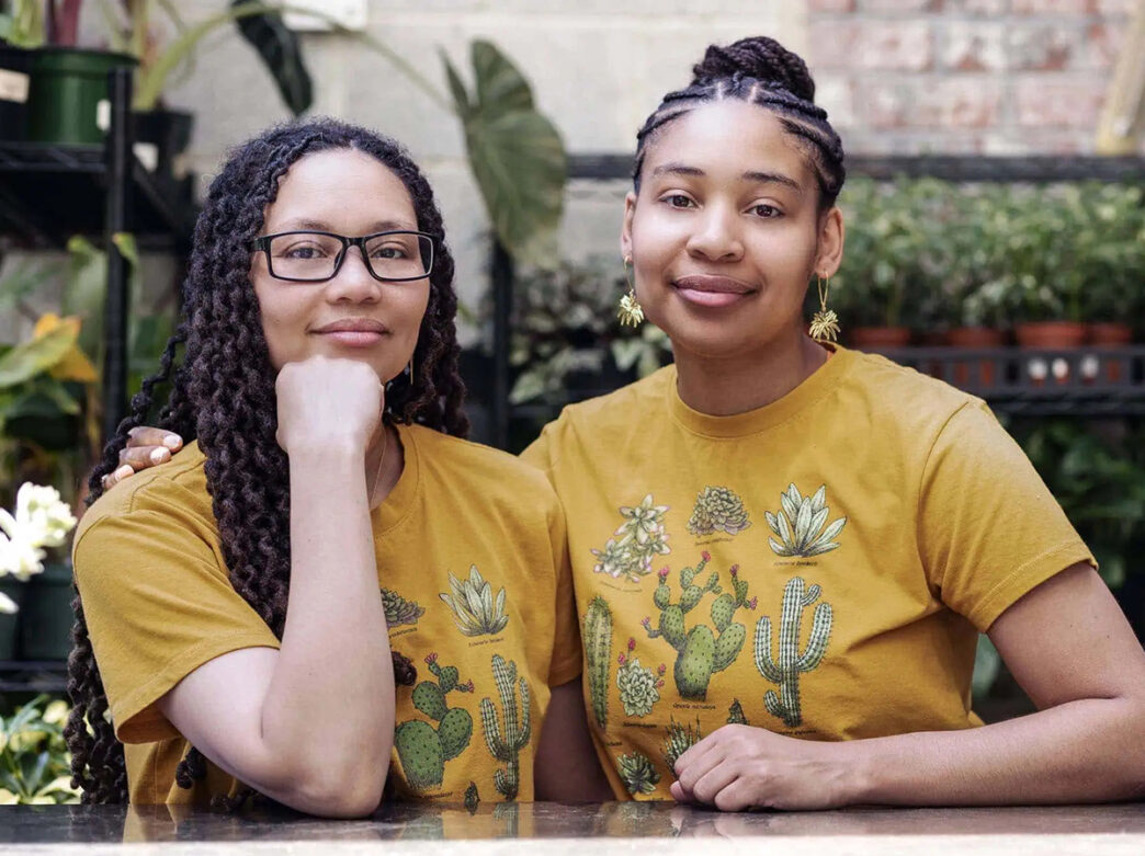 Diana and Breana, owners of Vault + Vine, pose for a photo together in front of green plants and a brick wall.
