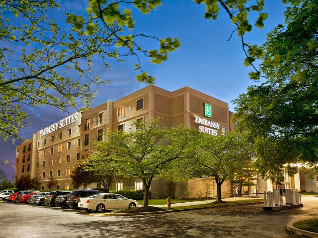 The Embassy Suites hotel at dusk, with its logo illuminated on the facade, and surrounded by trees and a parking area with cars.