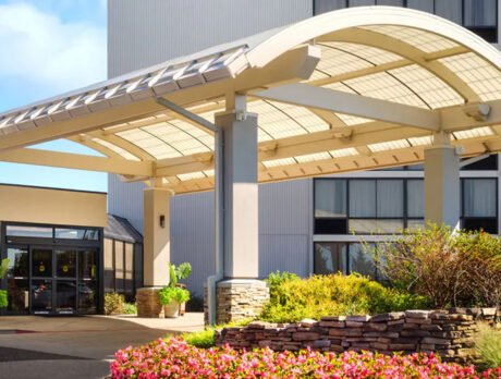 A welcoming hotel entrance featuring a modern arched canopy, a well-lit driveway, and attractive landscaping with colorful flowers and stone accents.