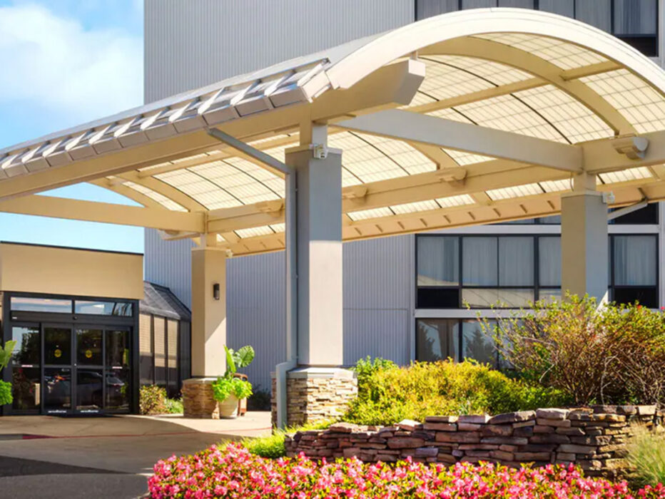 Une entrée d'hôtel accueillante avec un auvent moderne en arc de cercle, une allée bien éclairée et un aménagement paysager attrayant avec des fleurs colorées et des accents en pierre.