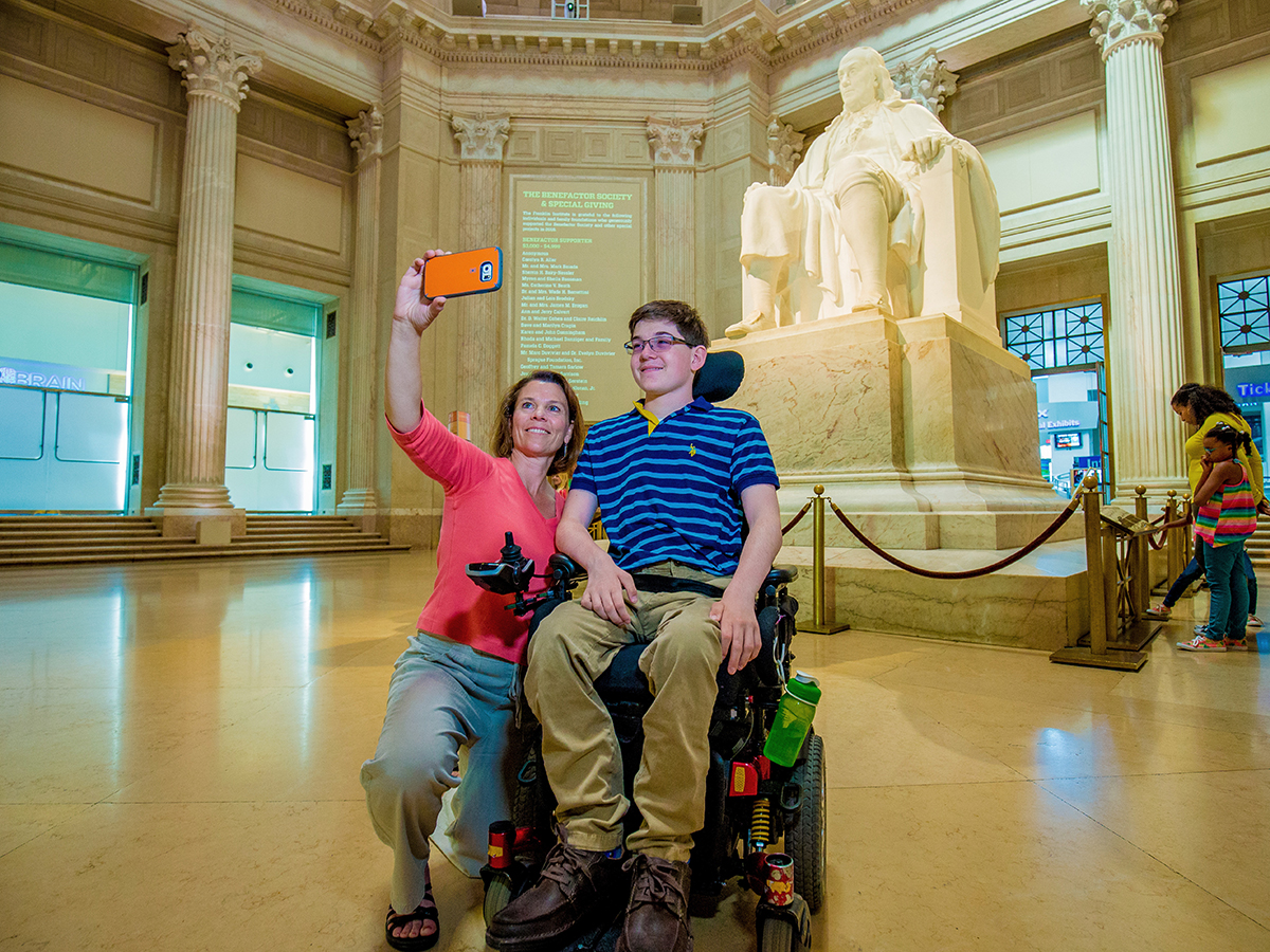 Two people take a selfie together in front of the Benjamin Franklin marble statue at the Franklin Institute. One person utilizes a motorized wheelchair.