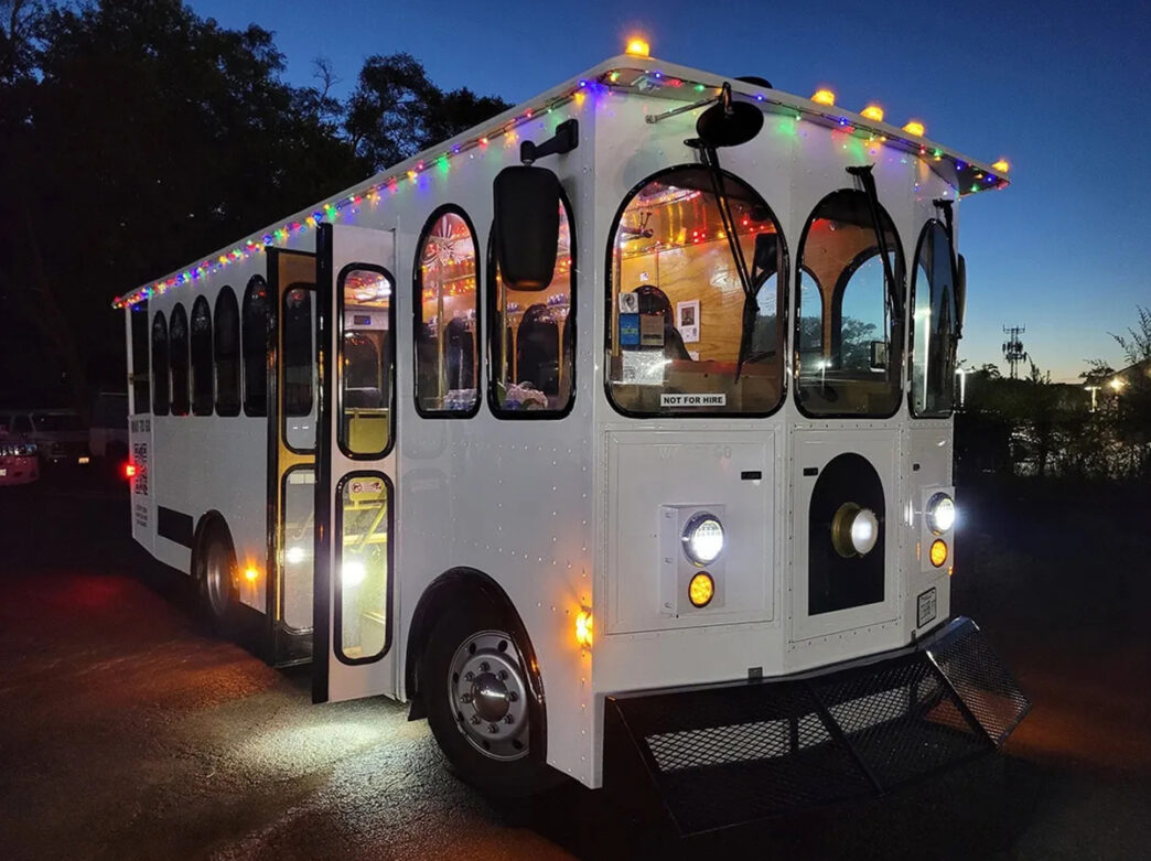 A white trolley is decorated with colorful string lights for the Holiday Lights Tour by Philadelphia Sightseeing Tours. The trolley is parked against a dark sky.