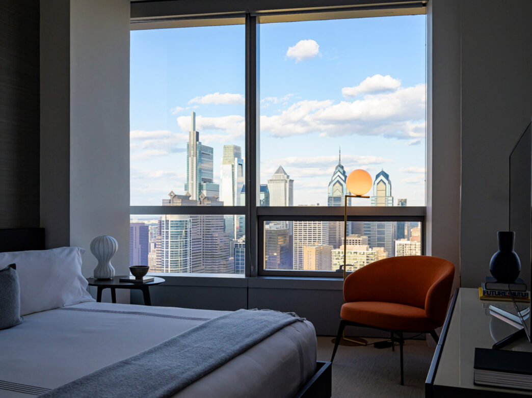 A modern hotel room inside Hotel AKA University City with a king-size bed and an orange chair facing a large window with a stunning view of the Philadelphia skyline.