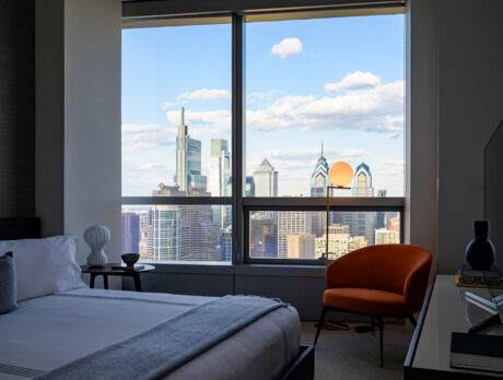 A modern hotel room inside Hotel AKA University City with a king-size bed and an orange chair facing a large window with a stunning view of the Philadelphia skyline.