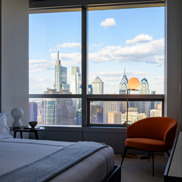 Una moderna habitación del Hotel AKA University City con una cama king size y una silla naranja frente a una gran ventana con impresionantes vistas al horizonte de Filadelfia.