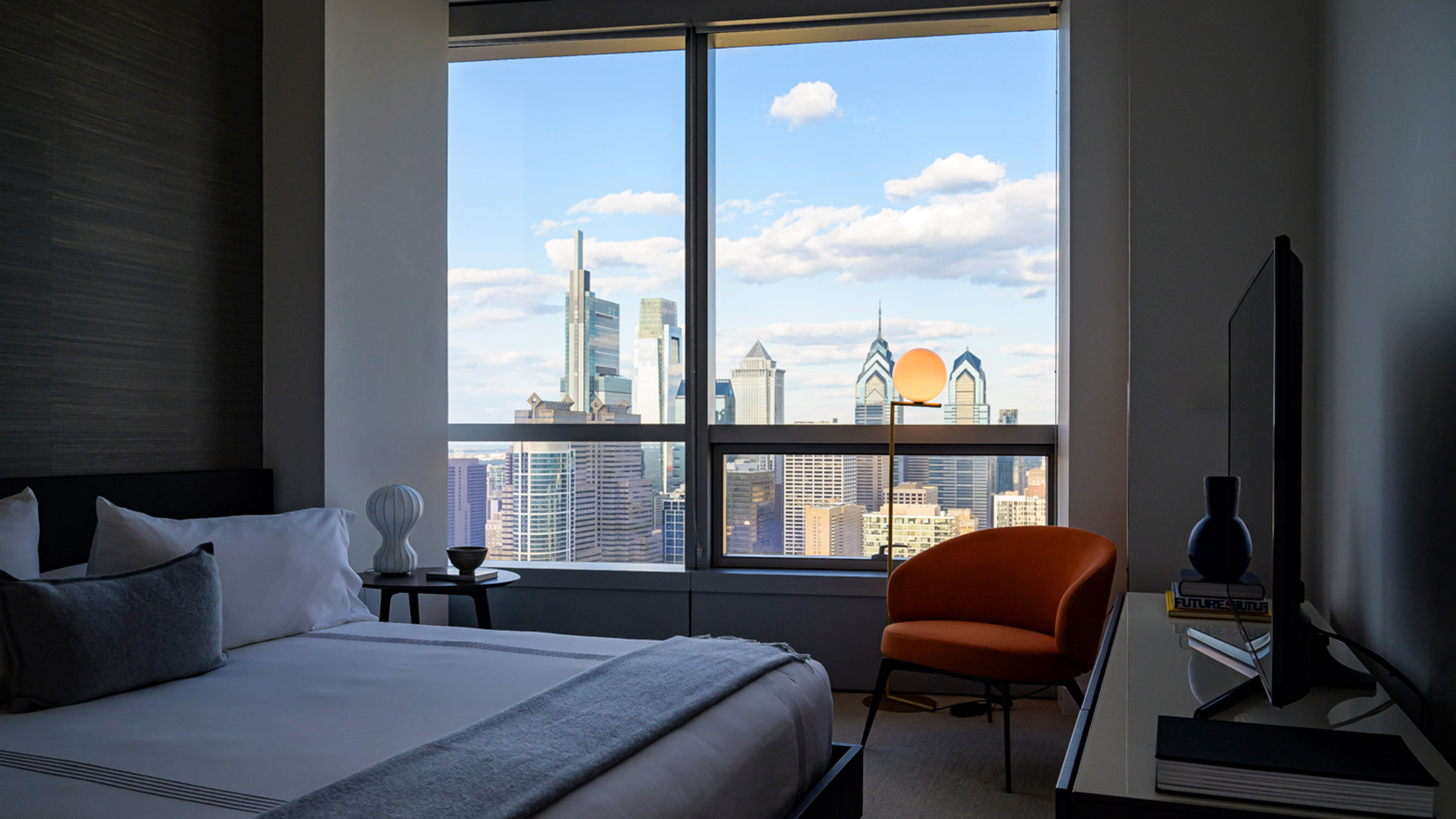 Um quarto de hotel moderno dentro do Hotel AKA University City com uma cama king-size e uma cadeira laranja de frente para uma grande janela com uma vista deslumbrante do horizonte da Filadélfia.