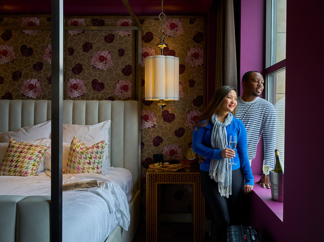 Een gedurfde gastenkamer in Hotel Monaco met een stijlvol grijs bed, bruin en bordeauxrood behang met bloemenpatroon, pruimkleurige muren en een raam.