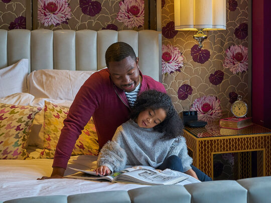 A bold guest room inside Hotel Monaco with tan and burgundy floral patterned wallpaper and a stylish gray bed. A father sits on a bed and reads with his daughter.