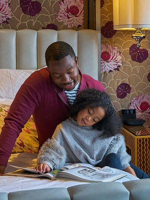 A bold guest room inside Hotel Monaco with tan and burgundy floral patterned wallpaper and a stylish gray bed. A father sits on a bed and reads with his daughter.