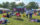A stage is set up in a grass lawn with white vendor tents on the perimeter for Indigenous Peoples Day Philly. People sit in lawn chairs and on the grass.