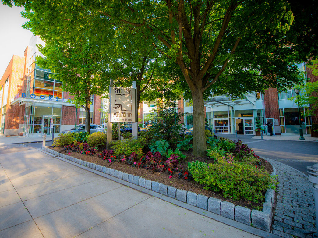 A lush green garden in front of the entrance of The Inn at Penn, a Hilton Hotel, on the University of Pennsylvania campus. A sign with the hotel's name is nestled within the garden.