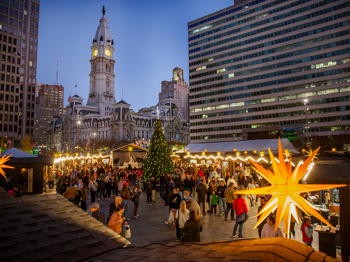 Vendor huts with decorative lighting and a large Christmas tree are set up in Love Park for Philadelphia's Christmas Village. People walk around shopping and taking photos. City Hall is in the background.