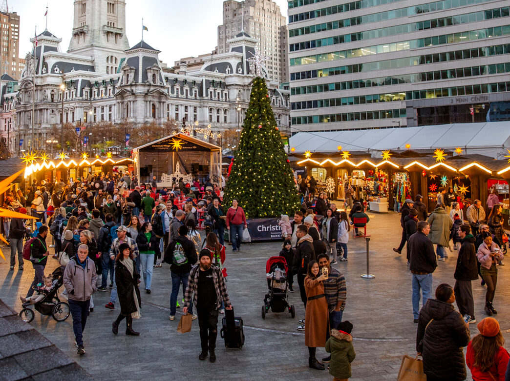 Vendor huts and a large Christmas tree are set up in Love Park for Philadelphia's Christmas Village. People walk around shopping and taking photos. City Hall is in the background.