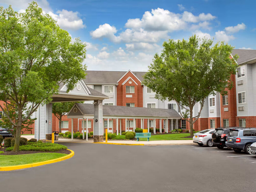 The gray and red brick facade of the Microtel Inn & Suites by Wyndham. The hotel building has a covered entrance and is surrounded by green trees and a parking area with cars.