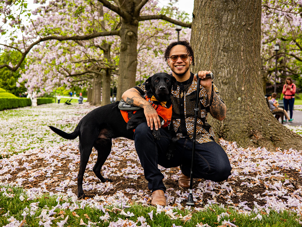 A person holding a cane kneels, smiles at the camera and wraps an arm around their service dog.
