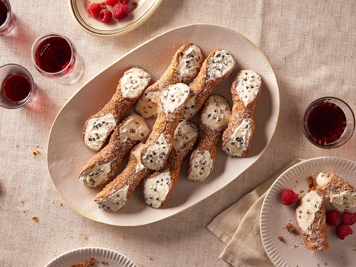 A platter of cannoli filled with ricotta, chocolate chips and cream. On the table there are also raspberries and glasses of red wine.