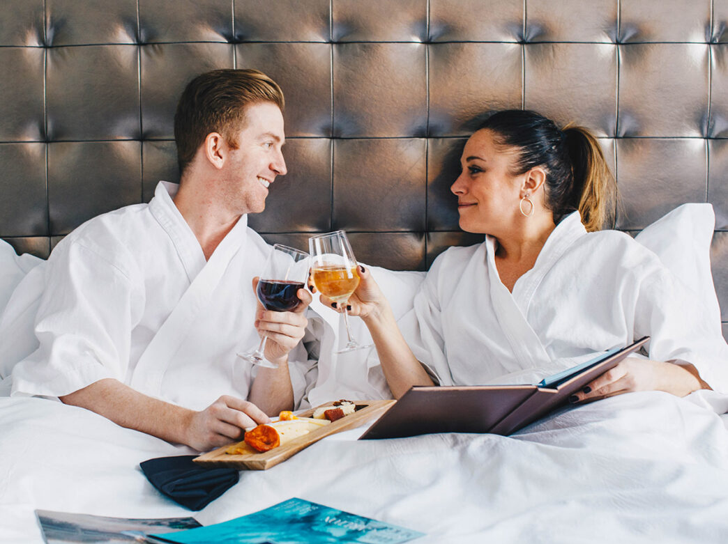 A couple, dressed in white linen robes, sit in bed and toast two glasses of wine while the gentleman holds a tray of charcuterie and the woman holds a brown book.