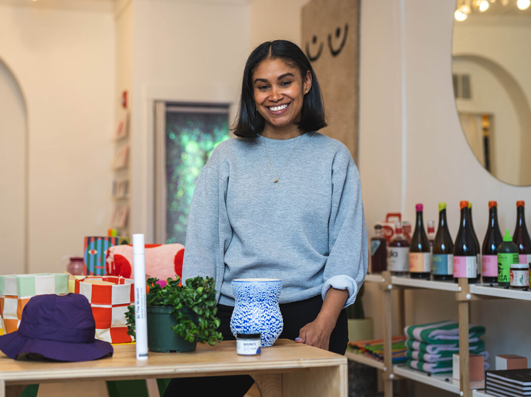 Shannon Maldonado, owner of Yowie, poses for a photo behind a table with homegoods and apparel. Behind her are shelves with bottles of wine and food items.