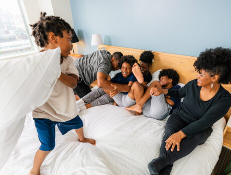 A happy, casually dressed family — mother, father and four children of various ages — laugh and play in a white bed at The Study hotel.