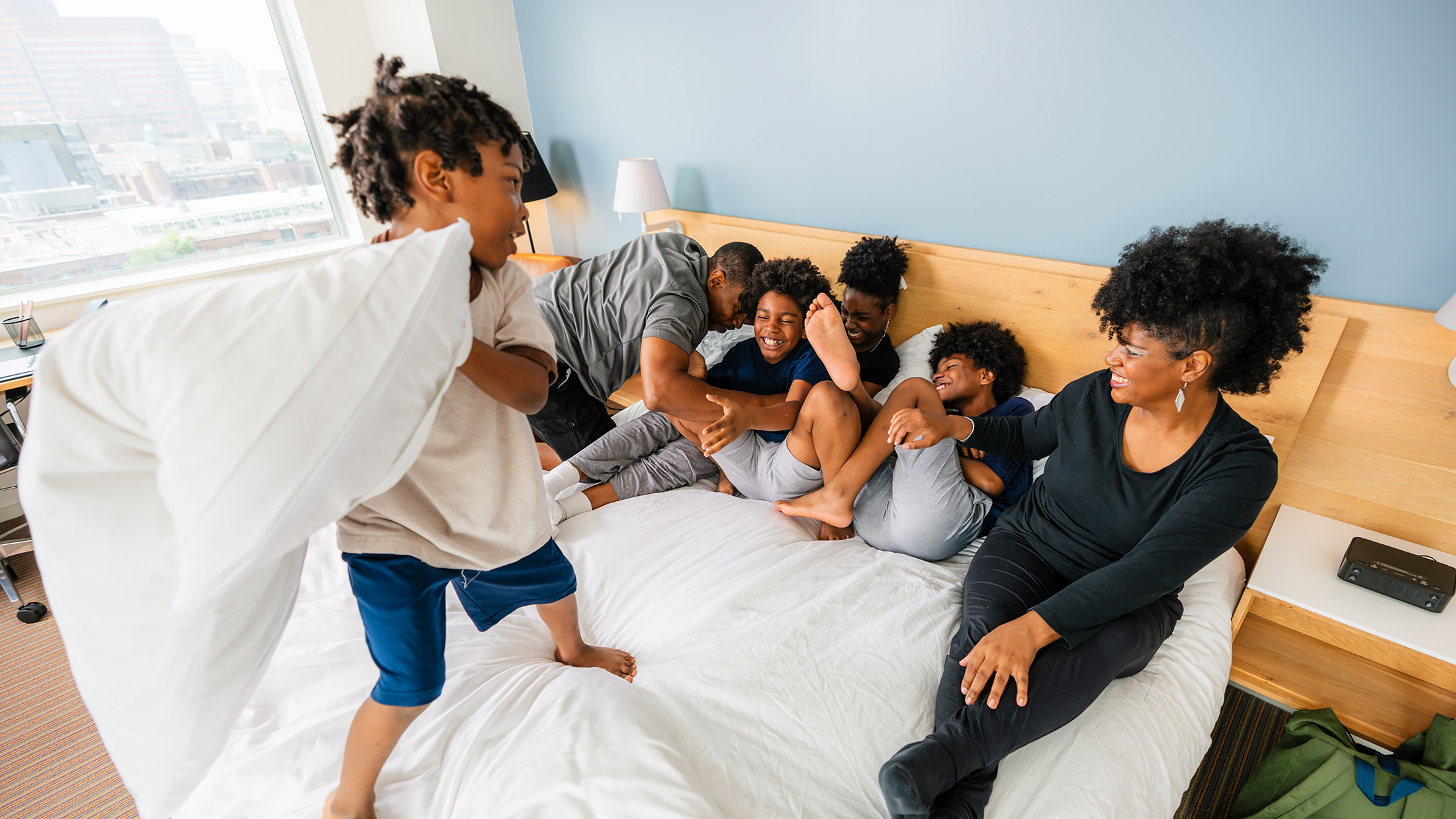 A happy, casually dressed family — mother, father and four children of various ages — laugh and play in a white bed at The Study hotel.