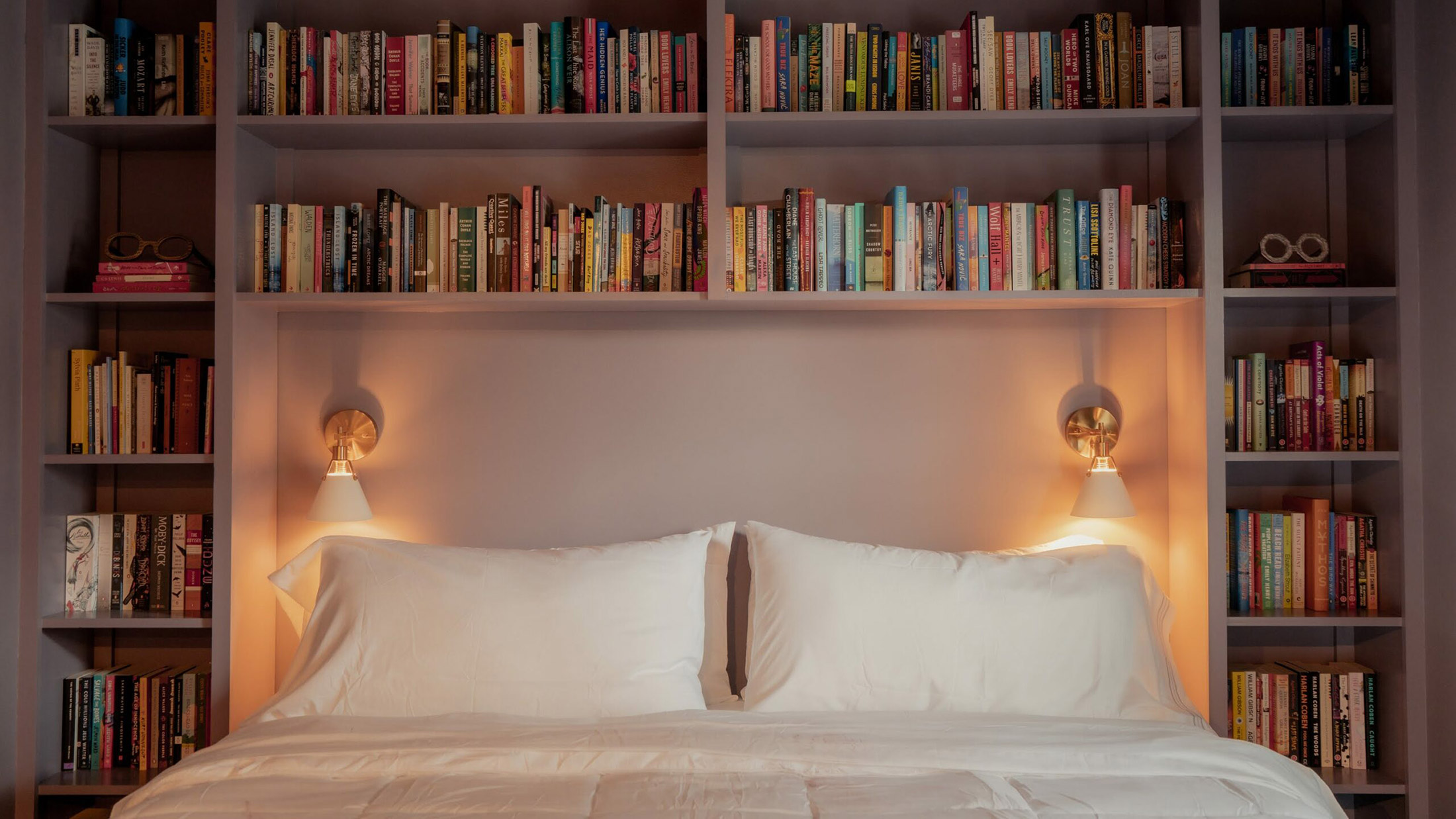 A bed with white sheets and pillows, flanked by built-in bookshelves and illuminated by wall-mounted reading lamps.