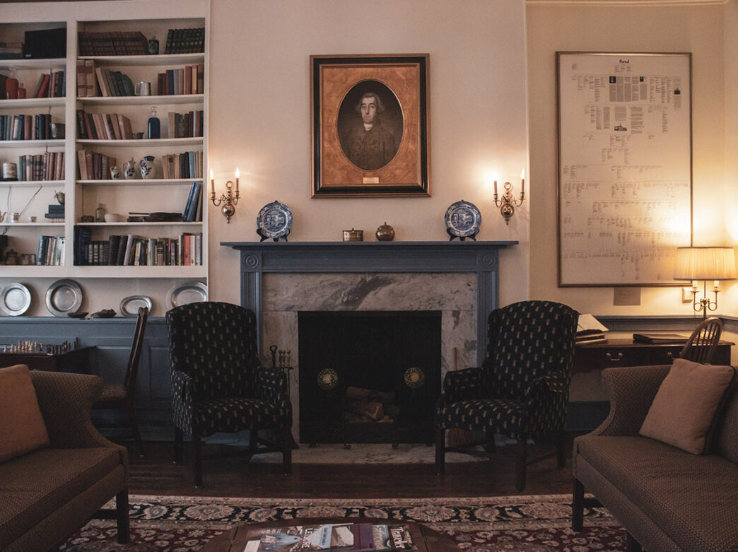 Een gezellige zithoek met een open haard, twee fauteuils, een bank en boekenplanken. Boven de open haard hangt een portret van een man uit de 18e eeuw en aan de rechtermuur hangt een wandtapijt.