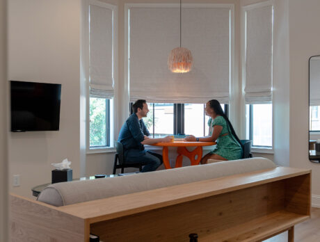 Surrounded by windows with the shades pulled halfway down, a couple sits at an orange table, under a tan fringe hanging light.