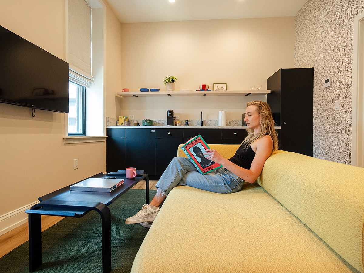 A woman dressed in a black top, blue jeans, and white sneakers sits on a pale yellow couch and reads the book "Afro-Atlantic Histories." A black coffee table, television, and black kitchen counter can also be seen in the room.