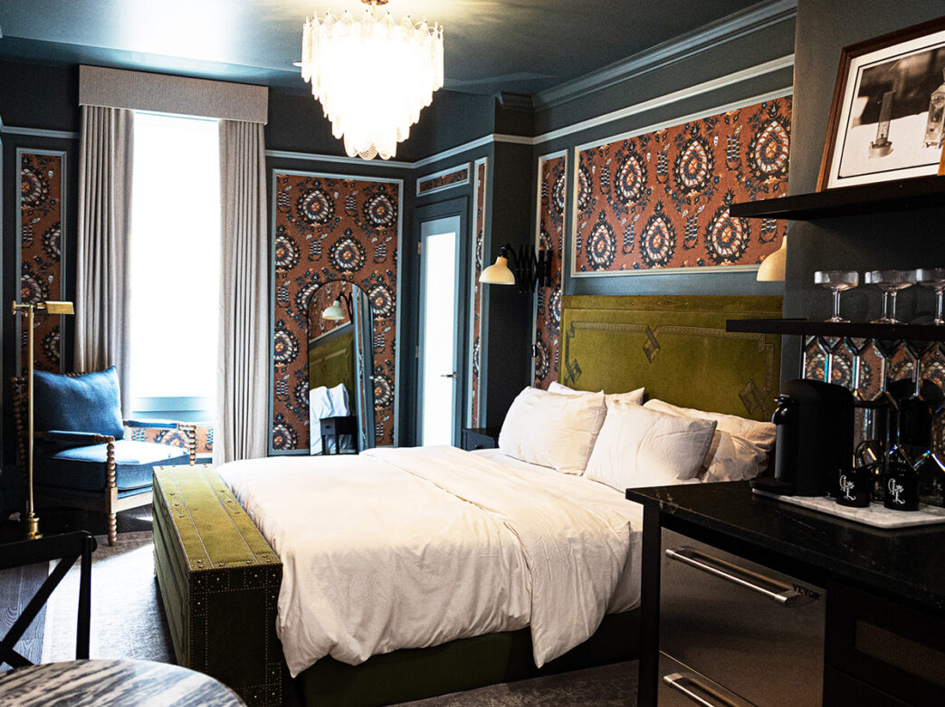 A hotel guestroom with a king bed, glass chandelier, mini bar and chair in the corner. The room is painted with a deep teal color and has wallpaper accent walls.