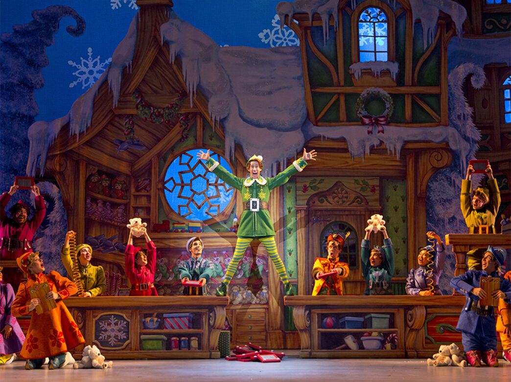 An actor dressed as Buddy the Elf stands on top of tables while other elves hold up Christmas toys during a production of Elf The Broadway Musical.