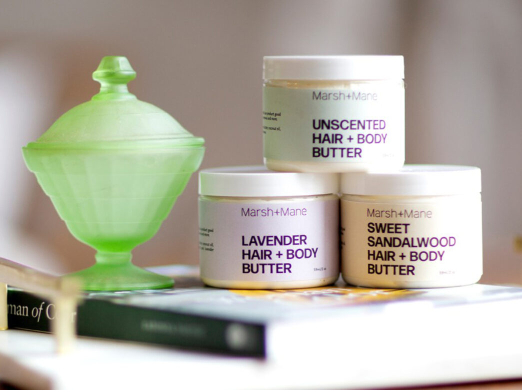 Three containers of hair and body butter are displayed next to a green decorative contrainer.