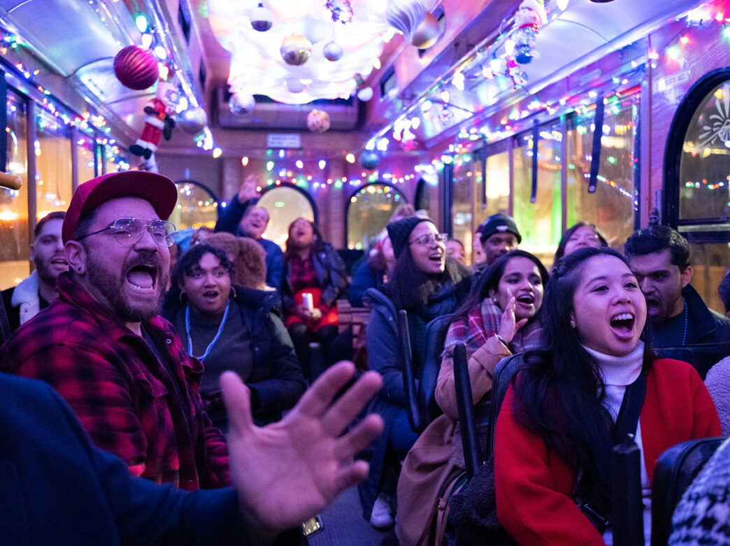 People cheer and sing while on the Holiday Light Trolley Tour. The inside of the trolley is decorated with colorful string lights and ornaments.