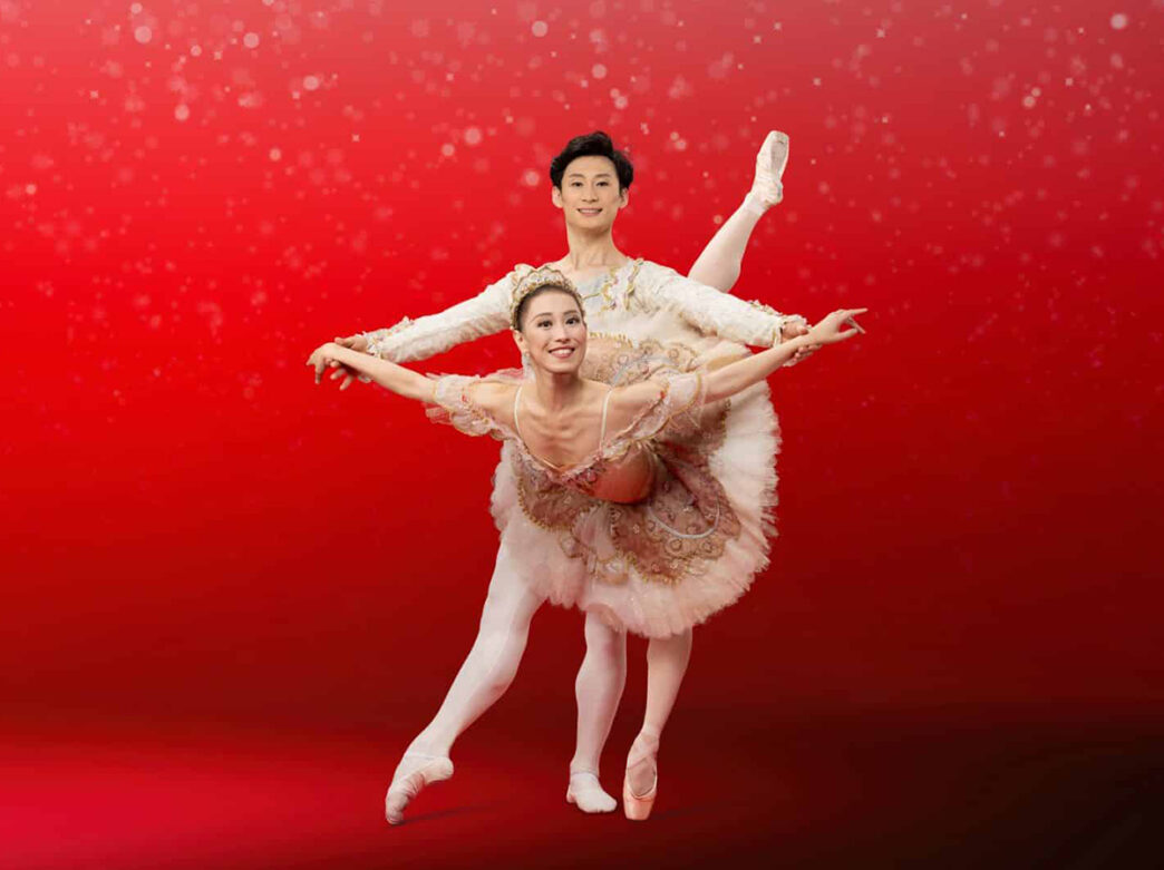 Two ballet dancers in The Nutcracker pose together while wearing white and gold costumes in front of a red background.