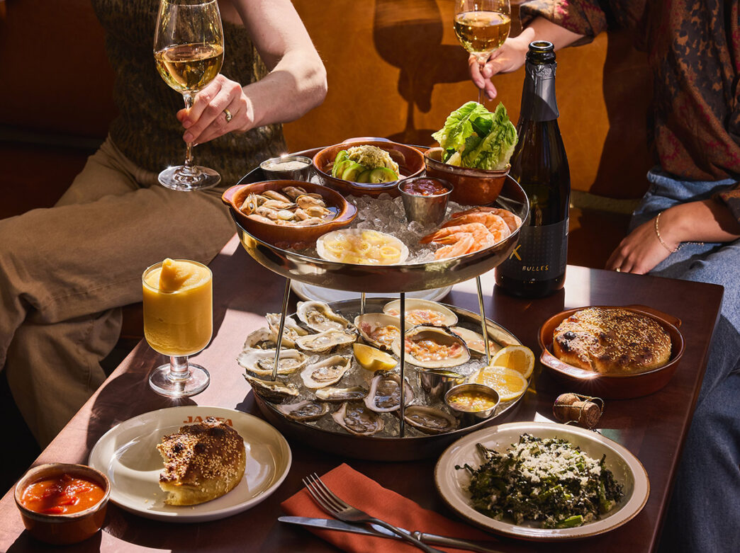 A two tier oyster tower with shrimp and clams is displayed in the center of a table. Other menu items surround the tower and people are seen in the background holding glasses of white wine.