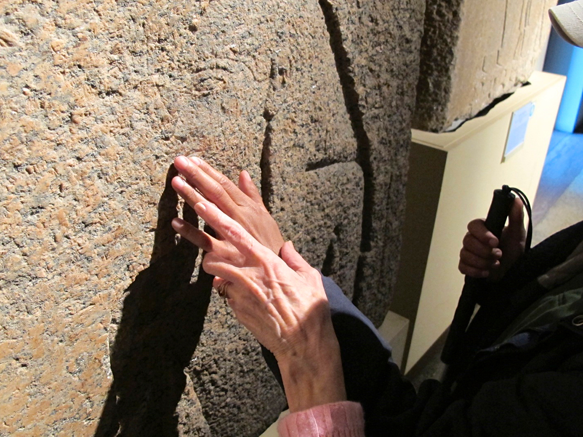 One person assists another person with touching an Egyptian hieroglyphic carved into granite.
