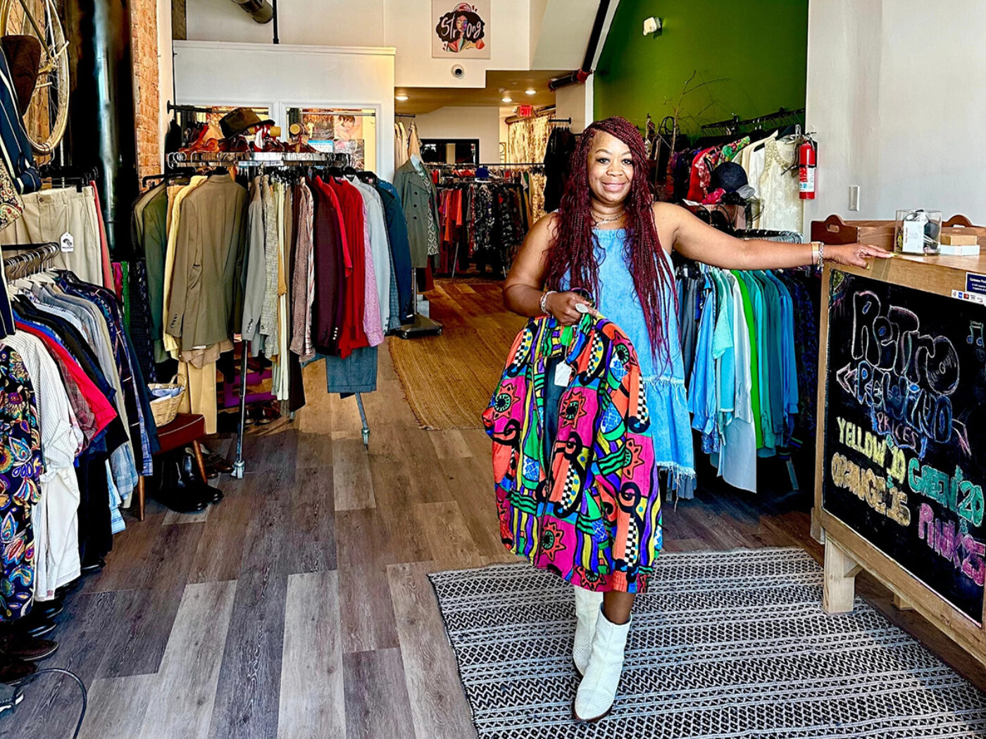 Tia Whitfield, owner of Retro Rewind Vintage and Trift, holds a colorful patterned jacket while smiling at the camera. The store is full of clothing racks and accessories.
