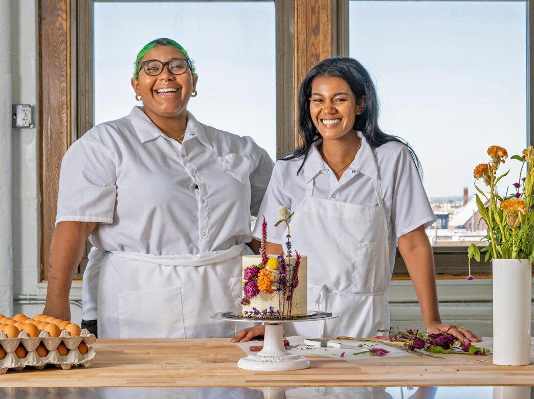 Two bakers wearing all white shirts and aprons stand behind a counter while smiling at the camera. On the counter is a small cake decorated with flowers.