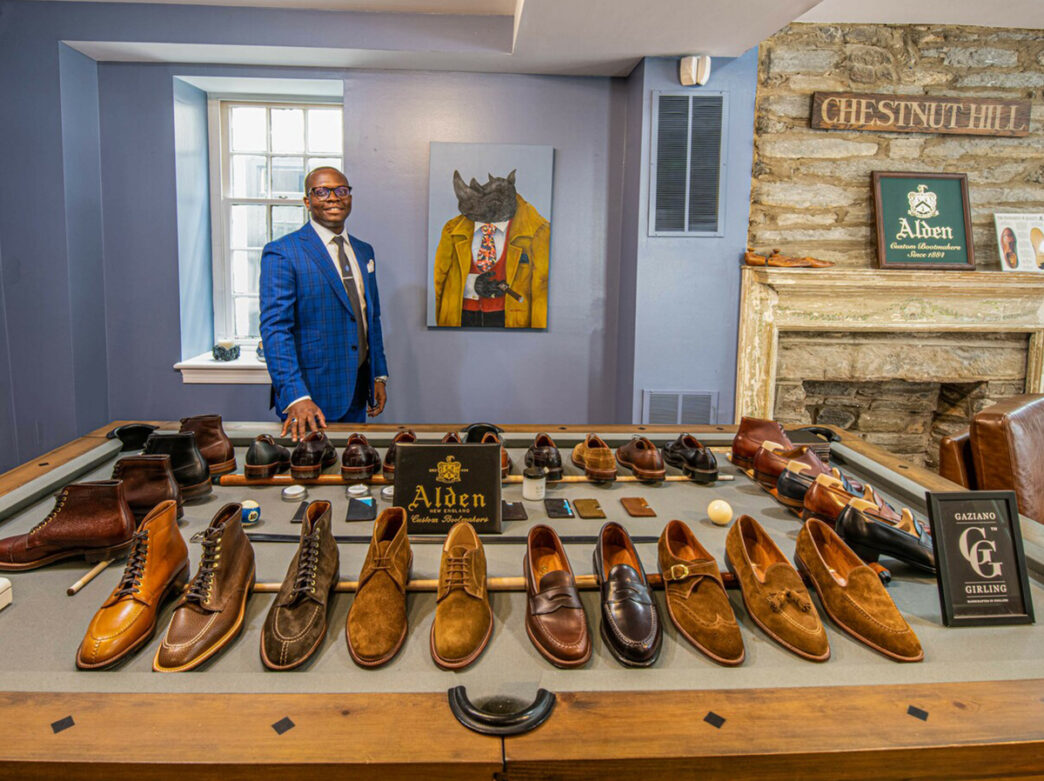 Voltaire Blain, owner of Style by Blain, wears a blue suit and stands behind a table with a display of shoes.