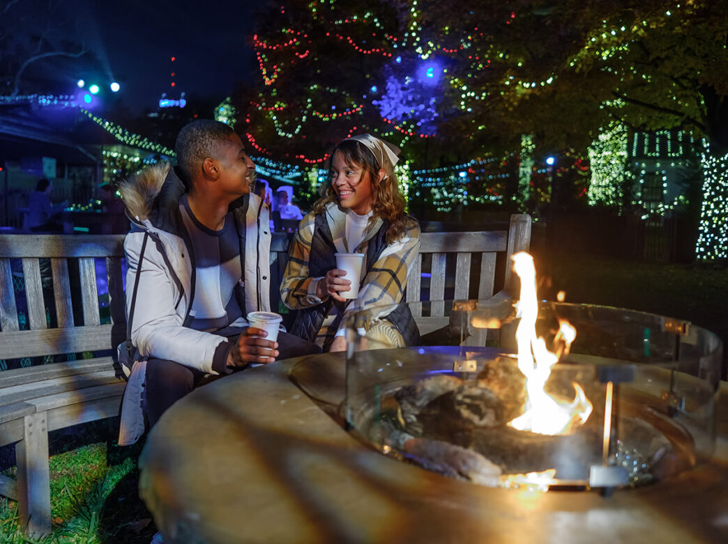 A couple sit on a wood bench behind a fire pit at Winter in Franklin Square. Both people hold cups and smile at each other.