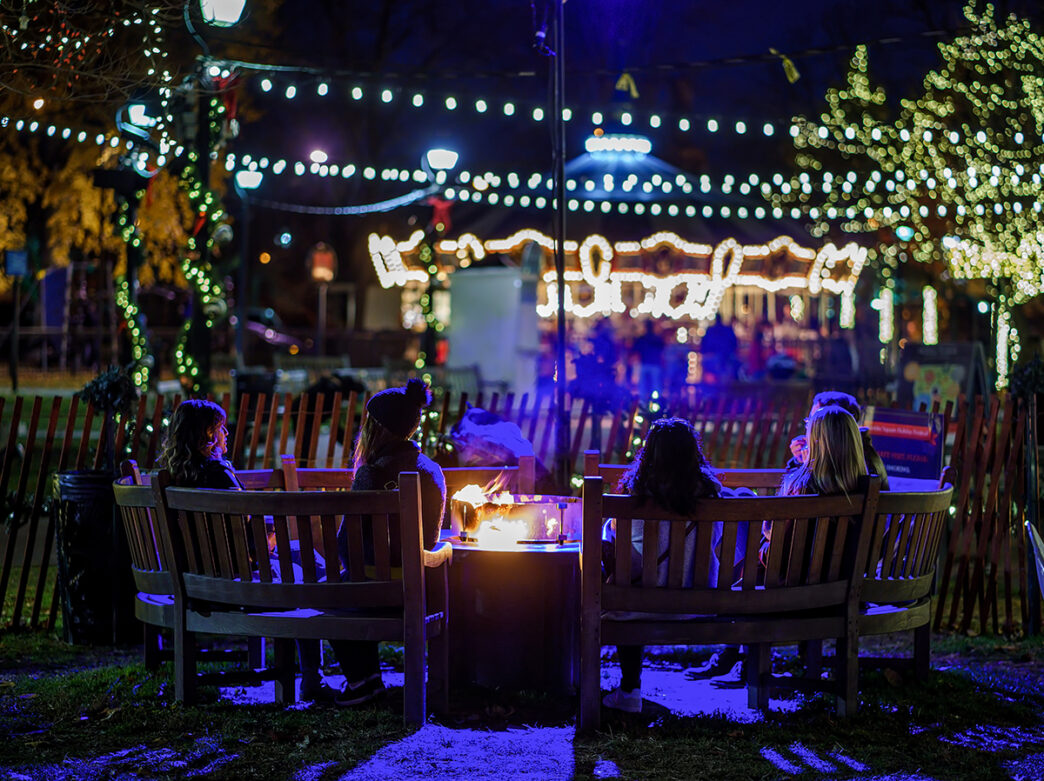 A group of people sit on benches around a fire pit at Franklin Square. String lights and a carousel is the background.
