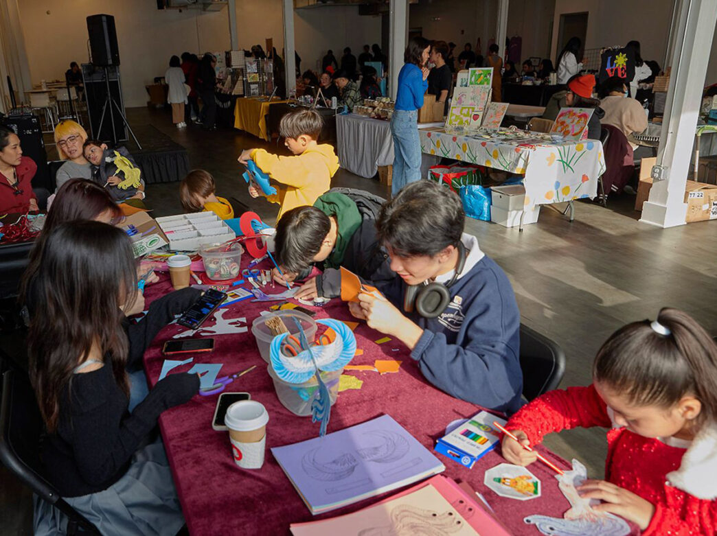 Kids sit around a table and do crafts while adults shop at vendor tables behind them at the Annual Community Fair.