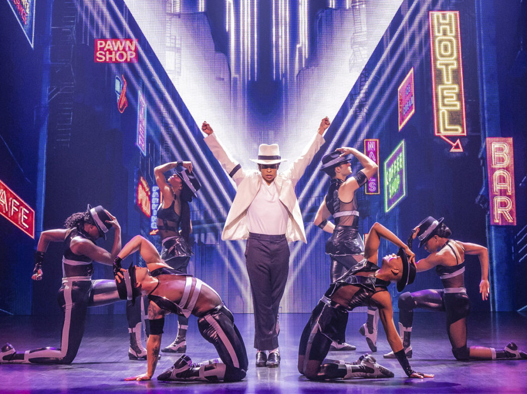 Set against a backdrop of glowing neon signs, the cast of MJ the Musical performs a high-energy dance number, with the lead actor in a white jacket and hat striking a pose surrounded by dancers in black-and-silver outfits.