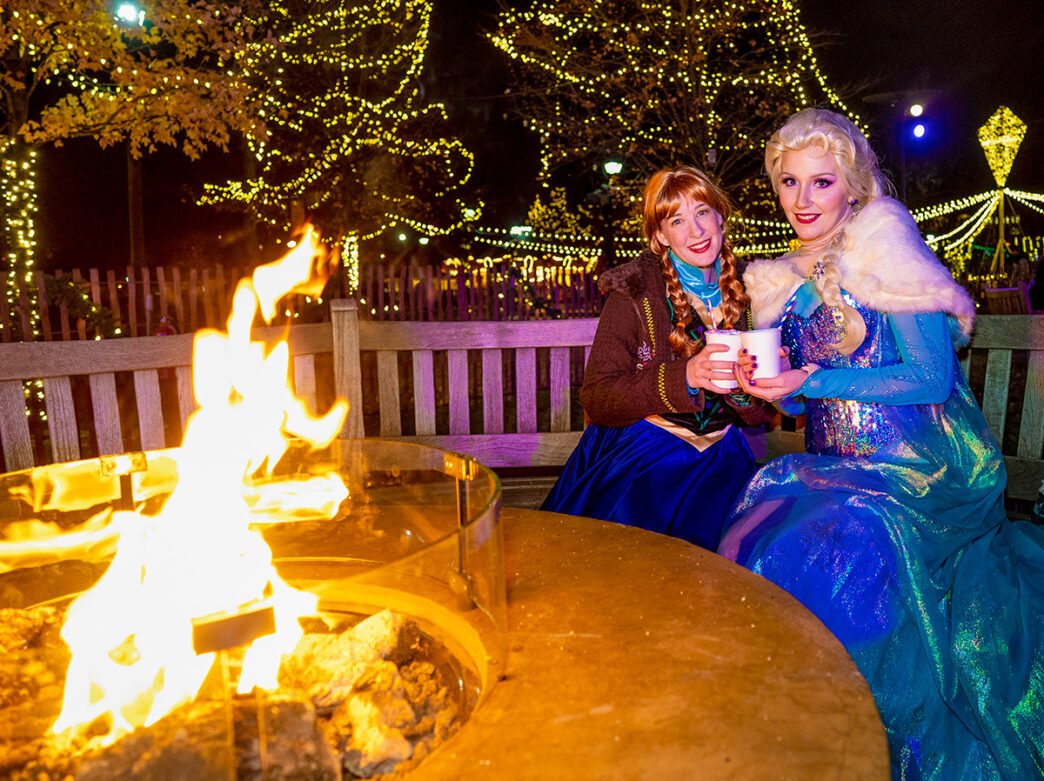 Surrounded by Franklin Square's sparkling lights, Anna and Elsa impersonators from Disney's Frozen sit by a cozy fire pit, holding warm drinks.