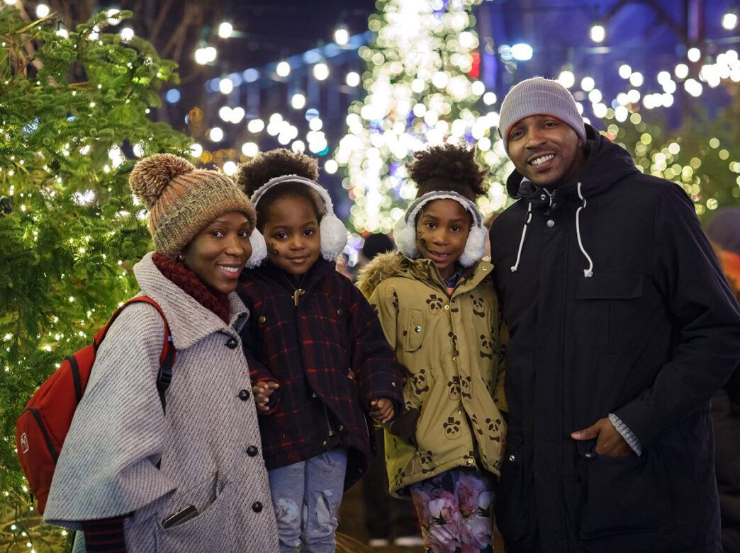 A smiling family of four poses among sparkling holiday lights and festive decorations at Independence Blue Cross Riverrink Winterfest.