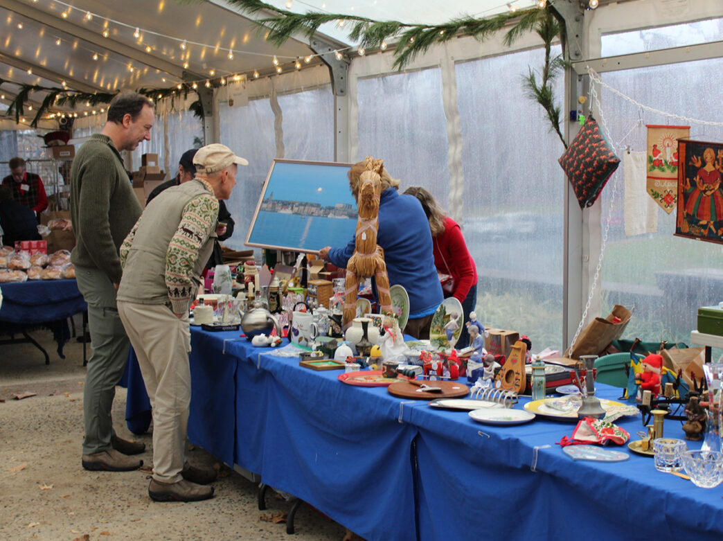 Two men look at a painting on display at the Lucia & Christmas Market at American Swedish Historical Museum. Other items for sale are displayed on a long table with a blue tablecloth.